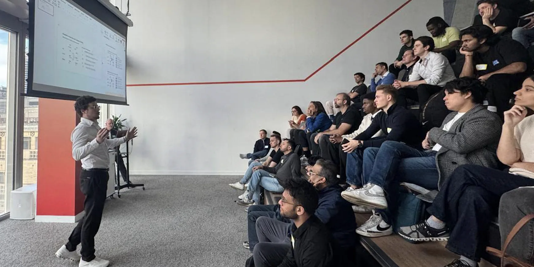 A speaker standing at the front of an office room, talking to about thirty people seated on tiered benches, with a spreadsheet on the projector screen behind him